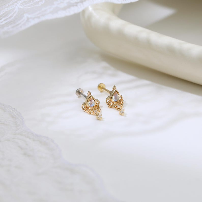 May include: Two gold-colored earrings with a teardrop-shaped iridescent stone and dangling chain details. The earrings have a post-back closure. The jewelry is displayed on a white surface.