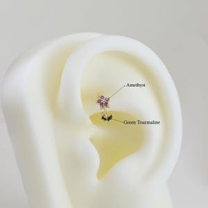 May include: A gold-colored earring with a floral design. The earring features a cluster of small purple amethyst stones and two green tourmaline leaves. The text "Amethyst" and "Green Tourmaline" are also visible.