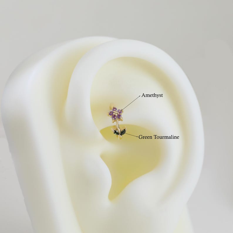 May include: A gold-colored earring with a floral design. The earring features a cluster of small purple amethyst stones and two green tourmaline leaves. The text "Amethyst" and "Green Tourmaline" are also visible.