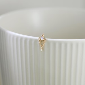 May include: A delicate gold-colored earring with a teardrop-shaped moonstone and dangling chain design. The earring features small, sparkling accents and a single, dangling white gem. The earring is set against a white background.