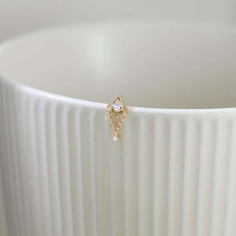 May include: A delicate gold-colored earring with a teardrop-shaped moonstone and dangling chain design. The earring features small, sparkling accents and a single, dangling white gem. The earring is set against a white background.