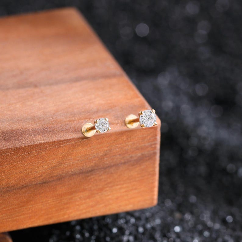Puede incluir: Dos pendientes de bot&oacute;n de oro, cada uno con una piedra preciosa redonda, transparente y facetada en un engaste de cuatro garras. Los pendientes tienen un cierre plano y circular y est&aacute;n colocados sobre una superficie de madera clara.