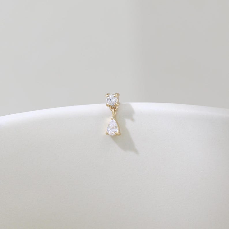 May include: A gold-colored earring with a square-cut clear gemstone at the top and a pear-shaped clear gemstone dangling below. The earring is displayed on a white surface.