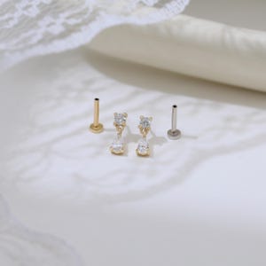May include: Gold and silver-toned body jewelry. Two gold-toned pieces feature a small, round top with a dangling teardrop-shaped gem. Two other pieces have a small, round top. The jewelry is displayed on a white surface.