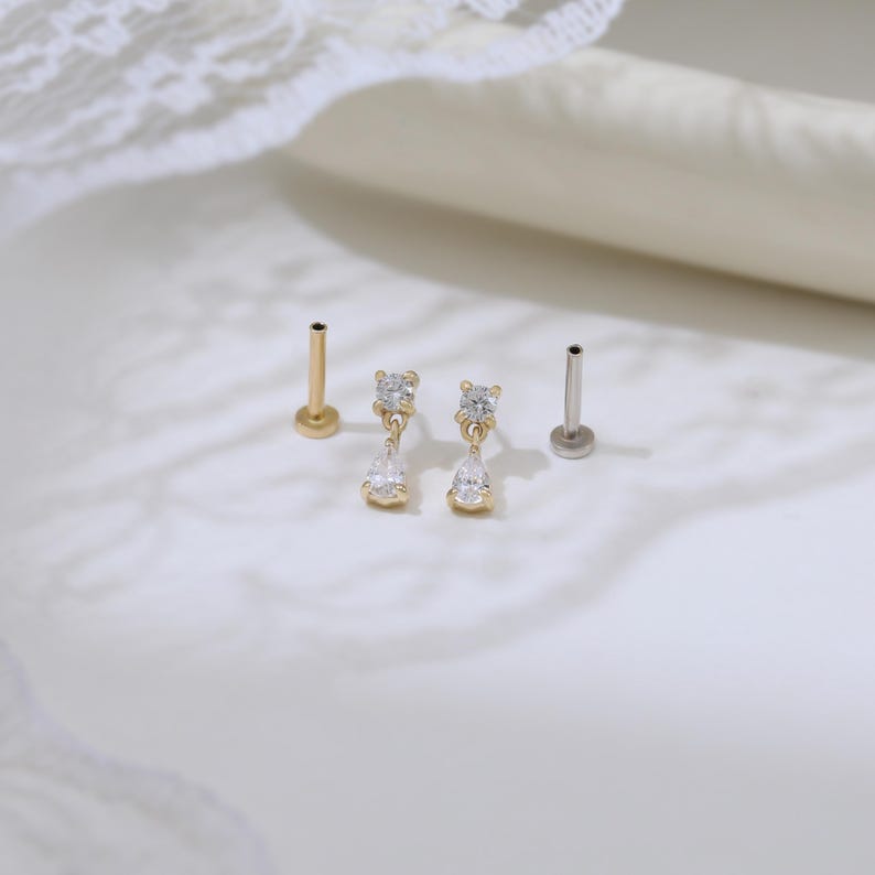 May include: Gold and silver-toned body jewelry. Two gold-toned pieces feature a small, round top with a dangling teardrop-shaped gem. Two other pieces have a small, round top. The jewelry is displayed on a white surface.
