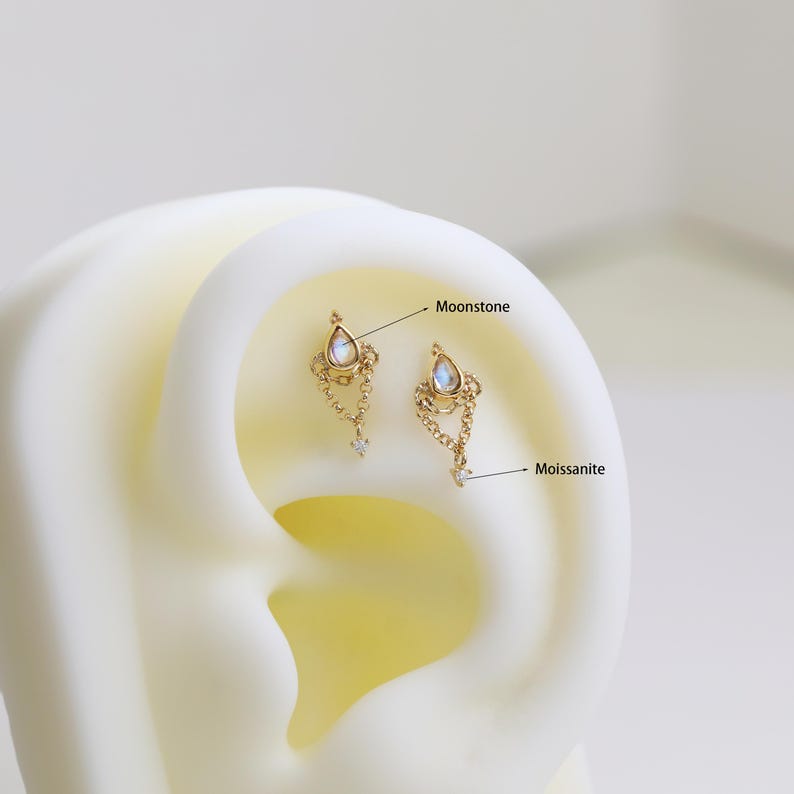 May include: Two gold-colored earrings with a teardrop-shaped moonstone and dangling chain design. Each earring has a small, clear moissanite stone at the bottom. The earrings are displayed on a white ear model.