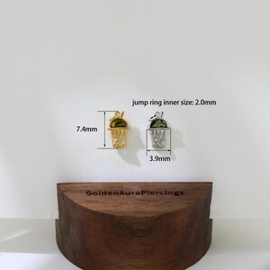 Peut inclure: Deux pendentifs de charme en or et en argent en forme de boisson glac&eacute;e verte avec une paille. Les pendentifs mesurent 7,4 mm et 3,9 mm. La taille int&eacute;rieure de l'anneau de saut est de 2,0 mm. Le texte "GoldenAuraPiercings" est visible sur la surface en bois.