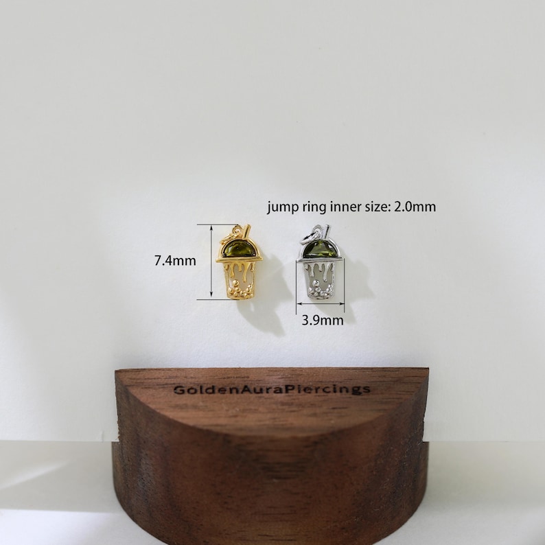 Peut inclure: Deux pendentifs de charme en or et en argent en forme de boisson glac&eacute;e verte avec une paille. Les pendentifs mesurent 7,4 mm et 3,9 mm. La taille int&eacute;rieure de l'anneau de saut est de 2,0 mm. Le texte "GoldenAuraPiercings" est visible sur la surface en bois.