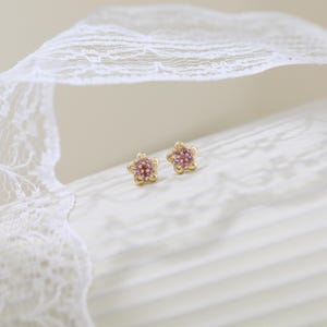 Pode incluir: Um par de brincos de ouro em forma de flores de cinco p&eacute;talas. Cada brinco apresenta um grupo de pequenas pedras preciosas redondas, cor-de-rosa e roxas, cravadas no centro das p&eacute;talas douradas, criando um design floral detalhado.