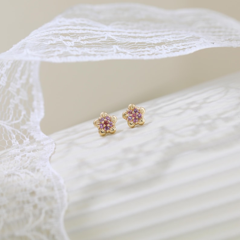 Pode incluir: Um par de brincos de ouro em forma de flores de cinco p&eacute;talas. Cada brinco apresenta um grupo de pequenas pedras preciosas redondas, cor-de-rosa e roxas, cravadas no centro das p&eacute;talas douradas, criando um design floral detalhado.