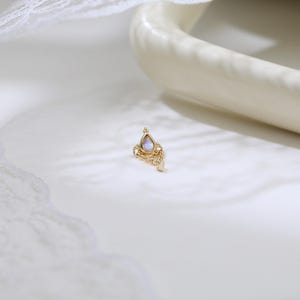 May include: A small, gold-colored earring with a teardrop-shaped, iridescent stone at the top. The earring has a decorative, filigree design with small, clear stones. The earring is set against a white background with a curved, off-white object.