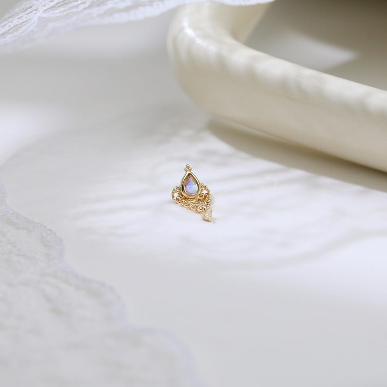 May include: A small, gold-colored earring with a teardrop-shaped, iridescent stone at the top. The earring has a decorative, filigree design with small, clear stones. The earring is set against a white background with a curved, off-white object.