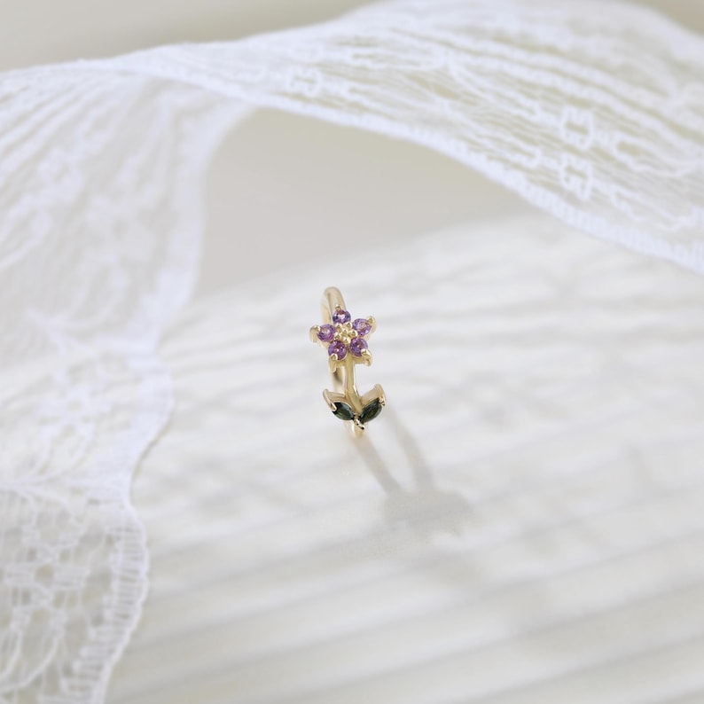 May include: A gold-colored floral ear cuff. The flower has purple gemstone petals and a yellow center. Two green leaves are at the base of the flower. The ear cuff is on a white surface.