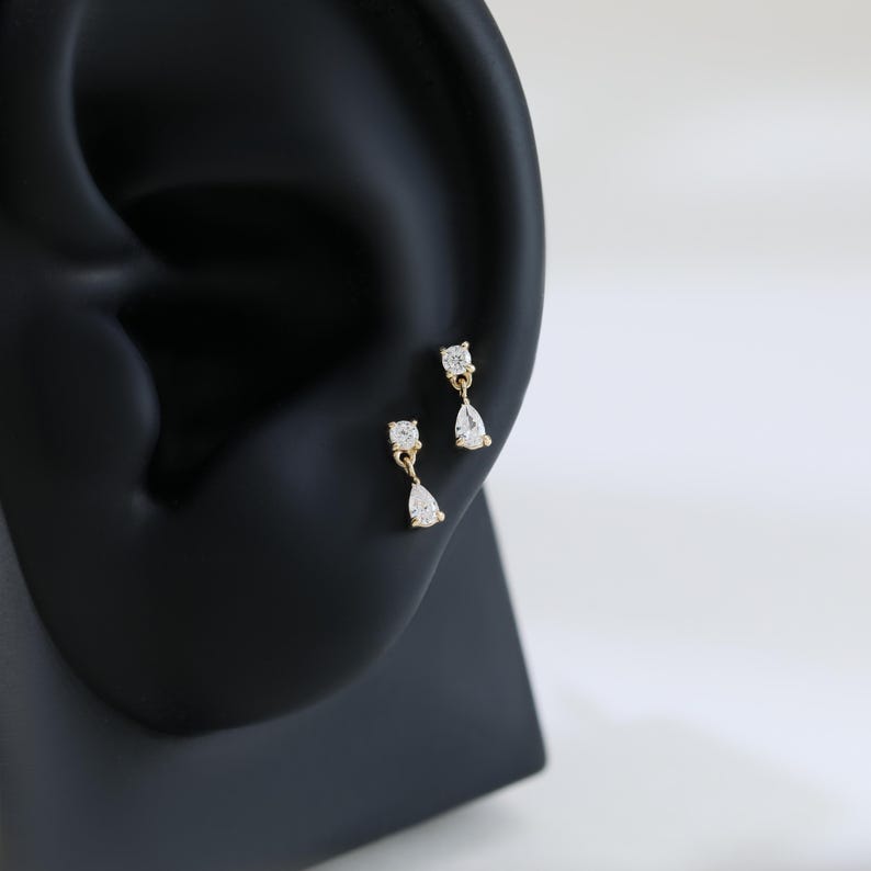 May include: A pair of gold-colored earrings with a square-cut and a teardrop-shaped clear stone. The earrings are displayed on a black ear-shaped form against a white background. The earrings are small and delicate.