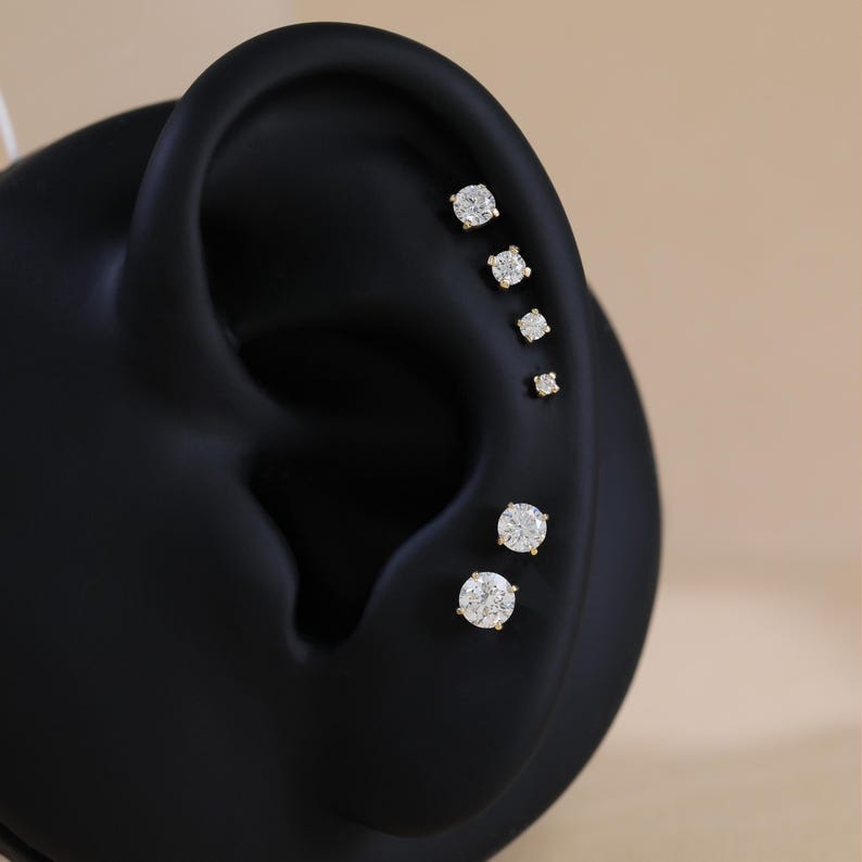 Puede incluir: Un conjunto de seis pendientes de bot&oacute;n redondos, transparentes y de talla brillante, dispuestos en una l&iacute;nea vertical a lo largo del borde exterior de un modelo de oreja negro. Cada pendiente tiene un engaste dorado de garras.