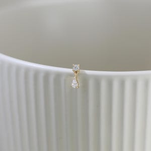 May include: A single gold earring with two clear, sparkling gemstones. The top stone is square, and the bottom stone is a teardrop shape. The earring is set against a white background.
