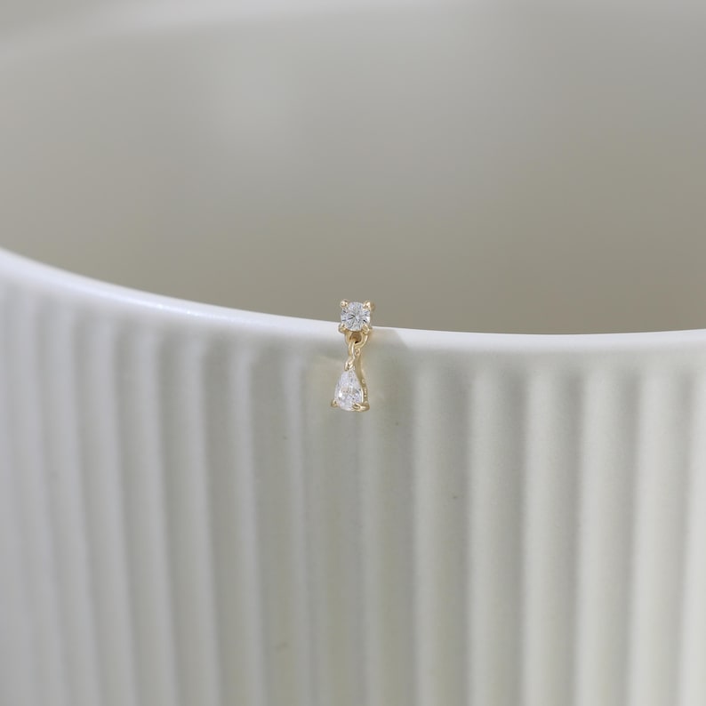 May include: A single gold earring with two clear, sparkling gemstones. The top stone is square, and the bottom stone is a teardrop shape. The earring is set against a white background.