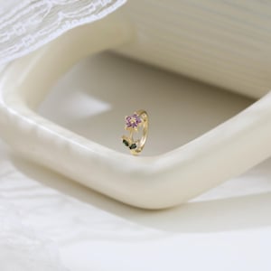 May include: A gold-colored ring featuring a flower design. The flower has purple and pink petals with green leaves. The ring is set against a white background.