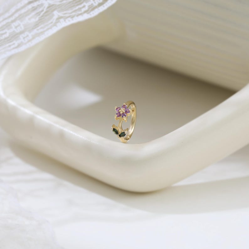 May include: A gold-colored ring featuring a flower design. The flower has purple and pink petals with green leaves. The ring is set against a white background.