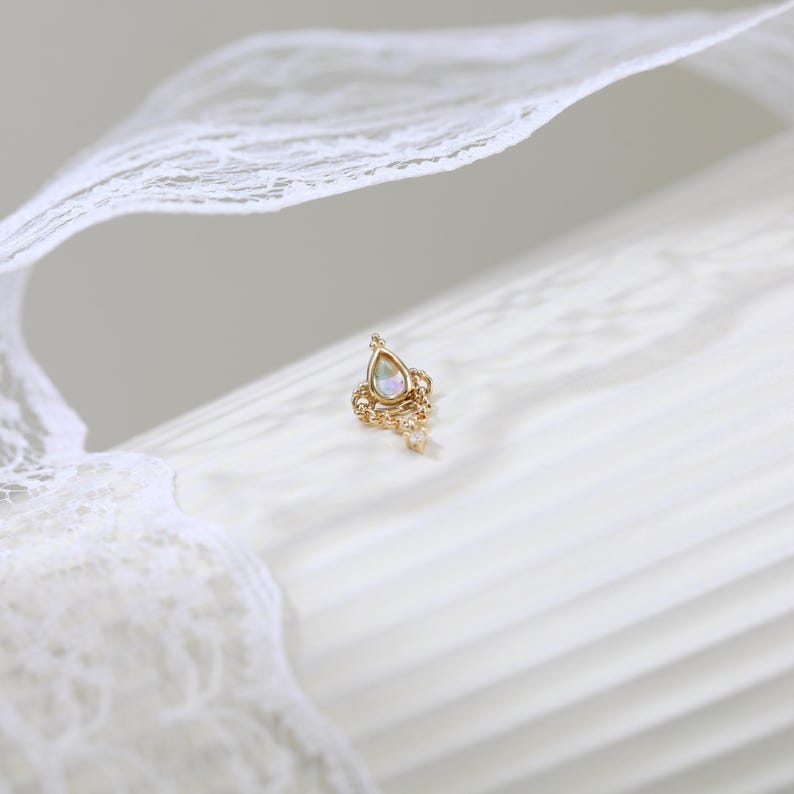 May include: A gold-colored earring with a teardrop-shaped opal stone and delicate chain details. The earring is set against a white background with a lace ribbon.