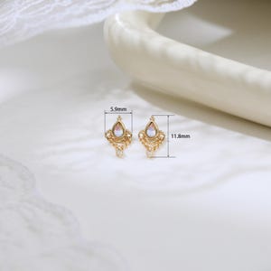 May include: A pair of gold-colored earrings with a pear-shaped, iridescent gemstone. Each earring is 0.46 inches long and 0.23 inches wide, featuring intricate filigree detailing and a small dangling accent. The earrings are displayed on a white surface.