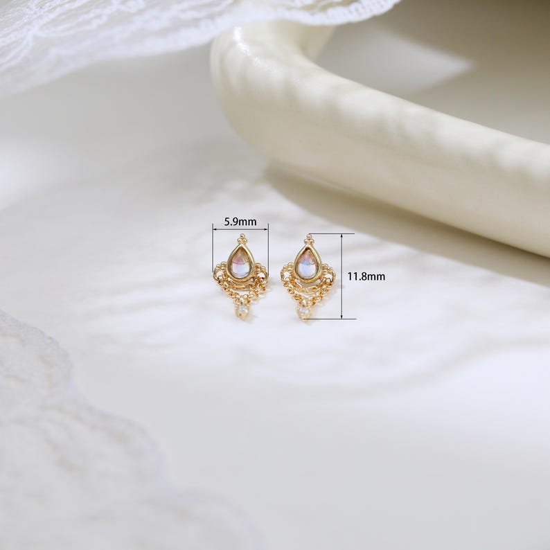 May include: A pair of gold-colored earrings with a pear-shaped, iridescent gemstone. Each earring is 0.46 inches long and 0.23 inches wide, featuring intricate filigree detailing and a small dangling accent. The earrings are displayed on a white surface.
