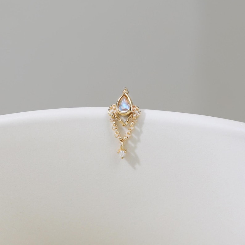 May include: A delicate gold-colored earring with a teardrop-shaped, iridescent gemstone at the top. The earring features a detailed filigree design and a small, dangling diamond-like accent at the bottom. The earring is set against a soft, neutral background.