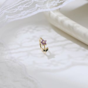 May include: A gold-colored earring with a floral design. The flower has purple gemstones at the center, with two dark green leaves. The earring is set against a white background with a lace detail.