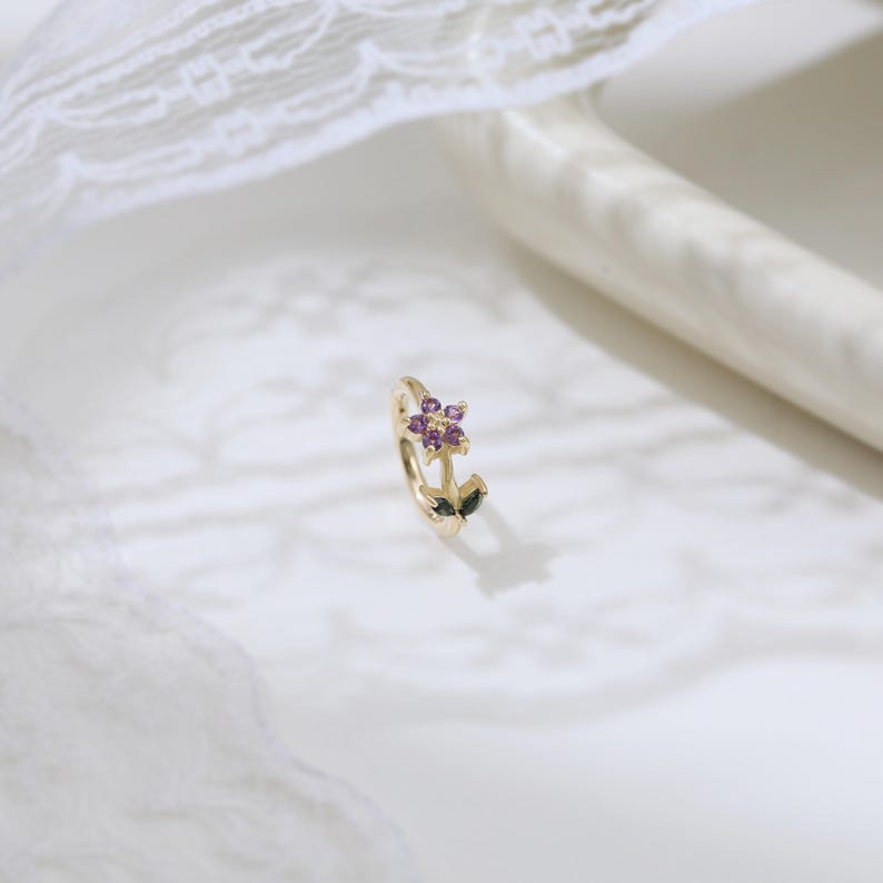 May include: A gold-colored earring with a floral design. The flower has purple gemstones at the center, with two dark green leaves. The earring is set against a white background with a lace detail.