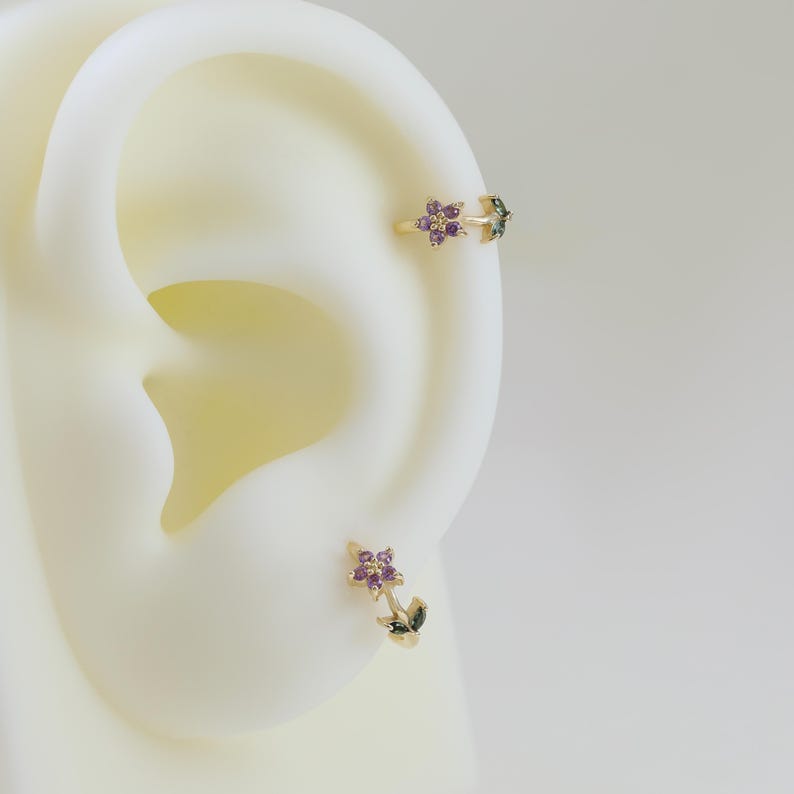 May include: Gold-toned ear jewelry featuring a floral design with purple and green gemstones. The set includes a cuff and a stud earring, both showcasing the flower motif. The jewelry is set against a neutral background, highlighting the intricate details.