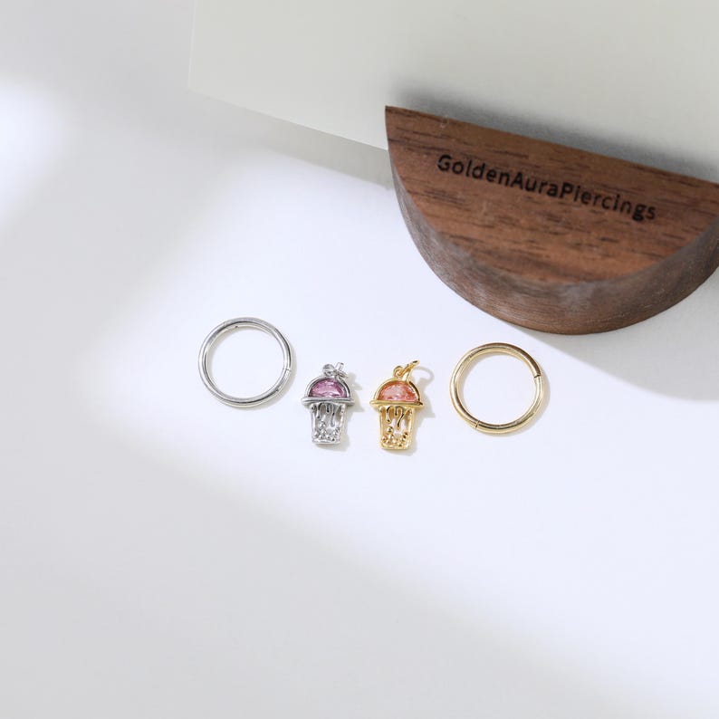 Peut inclure: Deux cr&eacute;oles dor&eacute;es avec un petit charme de ballon de basket attach&eacute; &agrave; chacune. Un charme a un ballon de basket rose et l'autre a un ballon de basket transparent. Les cr&eacute;oles sont sur une surface blanche avec un bloc de bois portant le texte "GoldenAuraPiercings".