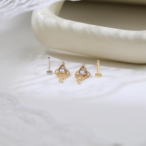 May include: Two gold-colored, ornate earrings with a teardrop-shaped, iridescent stone center. Each earring has a dangling charm and is accompanied by a post. The earrings are displayed on a white surface.