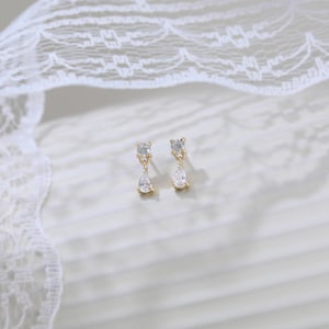May include: A pair of gold-toned drop earrings. Each earring features a small square-cut clear stone at the top, connected to a teardrop-shaped clear stone below. The earrings are set against a white background with a lace overlay.