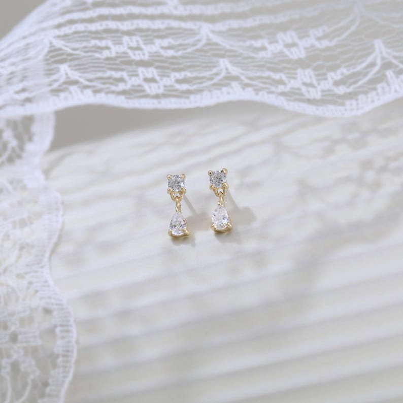 May include: A pair of gold-toned drop earrings. Each earring features a small square-cut clear stone at the top, connected to a teardrop-shaped clear stone below. The earrings are set against a white background with a lace overlay.