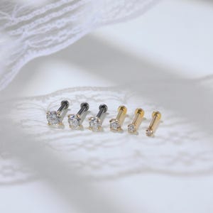 Puede incluir: Un conjunto de seis pendientes tipo labret dispuestos en fila. Tres pendientes tienen postes de metal gris oscuro con piedras redondas transparentes, mientras que los otros tres tienen postes de metal dorado con piedras redondas transparentes de varios tama&ntilde;os.