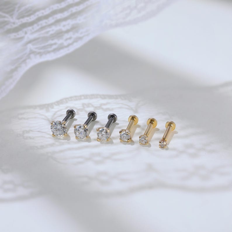 Puede incluir: Un conjunto de seis pendientes tipo labret dispuestos en fila. Tres pendientes tienen postes de metal gris oscuro con piedras redondas transparentes, mientras que los otros tres tienen postes de metal dorado con piedras redondas transparentes de varios tama&ntilde;os.