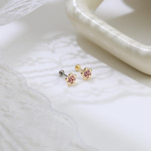 Pode incluir: Dois brincos de tarracha com um engaste dourado em forma de flor e um grupo de pequenas pedras cor-de-rosa no centro. Cada brinco tem um pino met&aacute;lico reto e um fecho circular plano para uma fixa&ccedil;&atilde;o segura no furo da orelha.