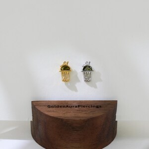 Peut inclure: Deux pendentifs de charme en or et en argent en forme de gobelet &agrave; boba avec une pierre pr&eacute;cieuse verte sur le dessus. Les breloques sont expos&eacute;es sur un support en bois avec le texte "GoldenAuraPiercings" sur le devant.