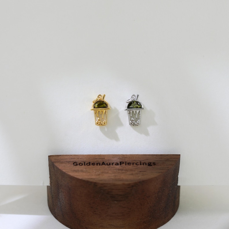 Peut inclure: Deux pendentifs de charme en or et en argent en forme de gobelet &agrave; boba avec une pierre pr&eacute;cieuse verte sur le dessus. Les breloques sont expos&eacute;es sur un support en bois avec le texte "GoldenAuraPiercings" sur le devant.