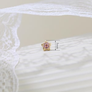 Pode incluir: Um brinco de ouro em forma de flor com cinco p&eacute;talas roxas claras e uma &uacute;nica pedra central vermelha. O brinco mede 5 mm de di&acirc;metro, conforme indicado pelas linhas de medi&ccedil;&atilde;o pretas e pelo texto vis&iacute;vel ao lado da pe&ccedil;a de joalharia.