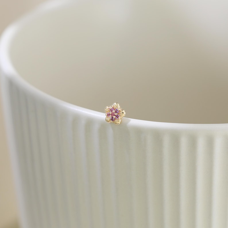 Pode incluir: Um pequeno brinco de ouro em forma de flor de cinco p&eacute;talas. O centro da flor apresenta um grupo de pequenas pedras preciosas redondas cor-de-rosa incrustadas na arma&ccedil;&atilde;o de metal dourado, criando um design floral delicado e detalhado.