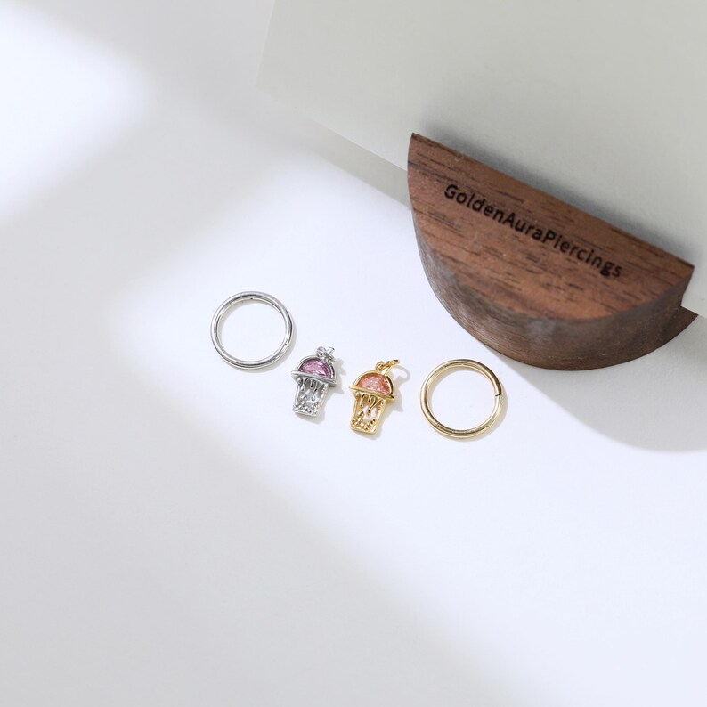 Peut inclure: Trois boucles d'oreilles en argent et en or avec un petit charme de montgolfi&egrave;re color&eacute; attach&eacute; &agrave; chacune. Les boucles d'oreilles sont pr&eacute;sent&eacute;es sur une surface en bois avec le texte "GoldenAuraPiercings" grav&eacute; dessus.