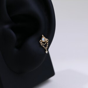 May include: A gold-colored earring with a teardrop-shaped gemstone and a dangling chain. The earring features a small, clear gemstone at the end of the chain. The earring is designed to be worn in a pierced ear.