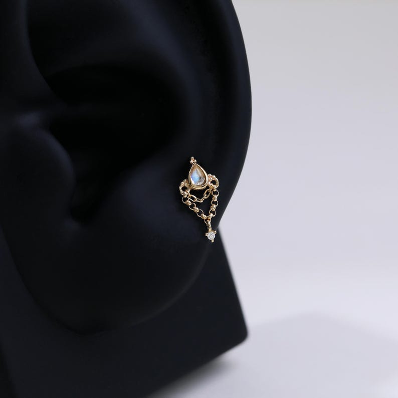 May include: A gold-colored earring with a teardrop-shaped gemstone and a dangling chain. The earring features a small, clear gemstone at the end of the chain. The earring is designed to be worn in a pierced ear.