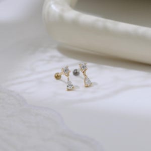 May include: Two gold-colored earrings with a post and a screw-on backing. Each earring features a square-cut clear stone at the top, with a pear-shaped clear stone dangling below. The earrings are displayed on a white surface.
