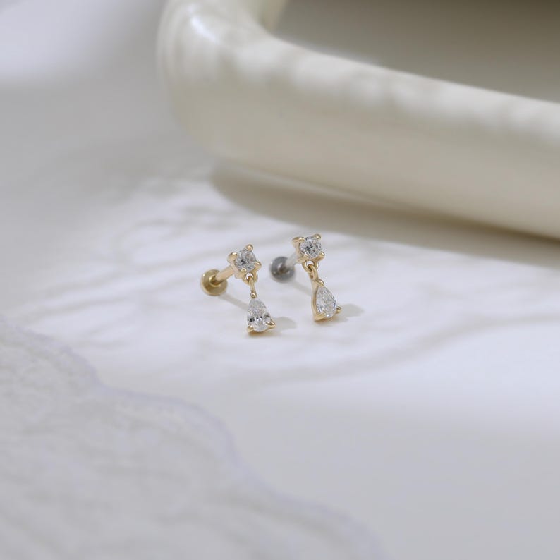 May include: Two gold-colored earrings with a post and a screw-on backing. Each earring features a square-cut clear stone at the top, with a pear-shaped clear stone dangling below. The earrings are displayed on a white surface.
