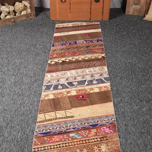 Puede incluir: Una alfombra pasillera larga y rectangular con un diseño de retazos de bandas horizontales en varios colores, incluyendo marrón, azul, naranja y crema. La alfombra muestra una mezcla de patrones geométricos, motivos florales y secciones lisas.