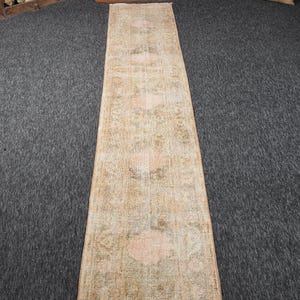 Può includere: Un tappeto lungo e stretto con un motivo sbiadito e usurato in tonalità tenui di beige, verde oliva e pesca delicato. Il tessuto rettangolare mostra un motivo geometrico e floreale ripetuto con un aspetto vintage invecchiato.