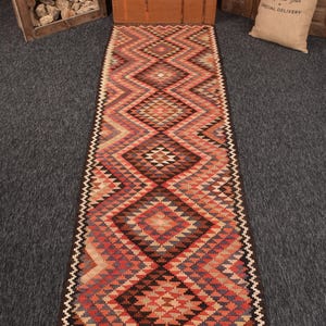 Puede incluir: Una alfombra larga y rectangular con un patrón geométrico de diamantes en tonos de rojo, marrón, beige y gris. La alfombra tiene un borde marrón oscuro y un borde en zigzag. Adecuada para un pasillo o entrada.