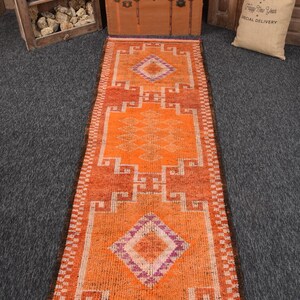 Puede incluir: Una alfombra de pasillo naranja y óxido con patrones geométricos. La alfombra presenta formas de diamante y rectangulares en tonos de naranja, óxido y morado. La alfombra es rectangular y tiene un borde con flecos.